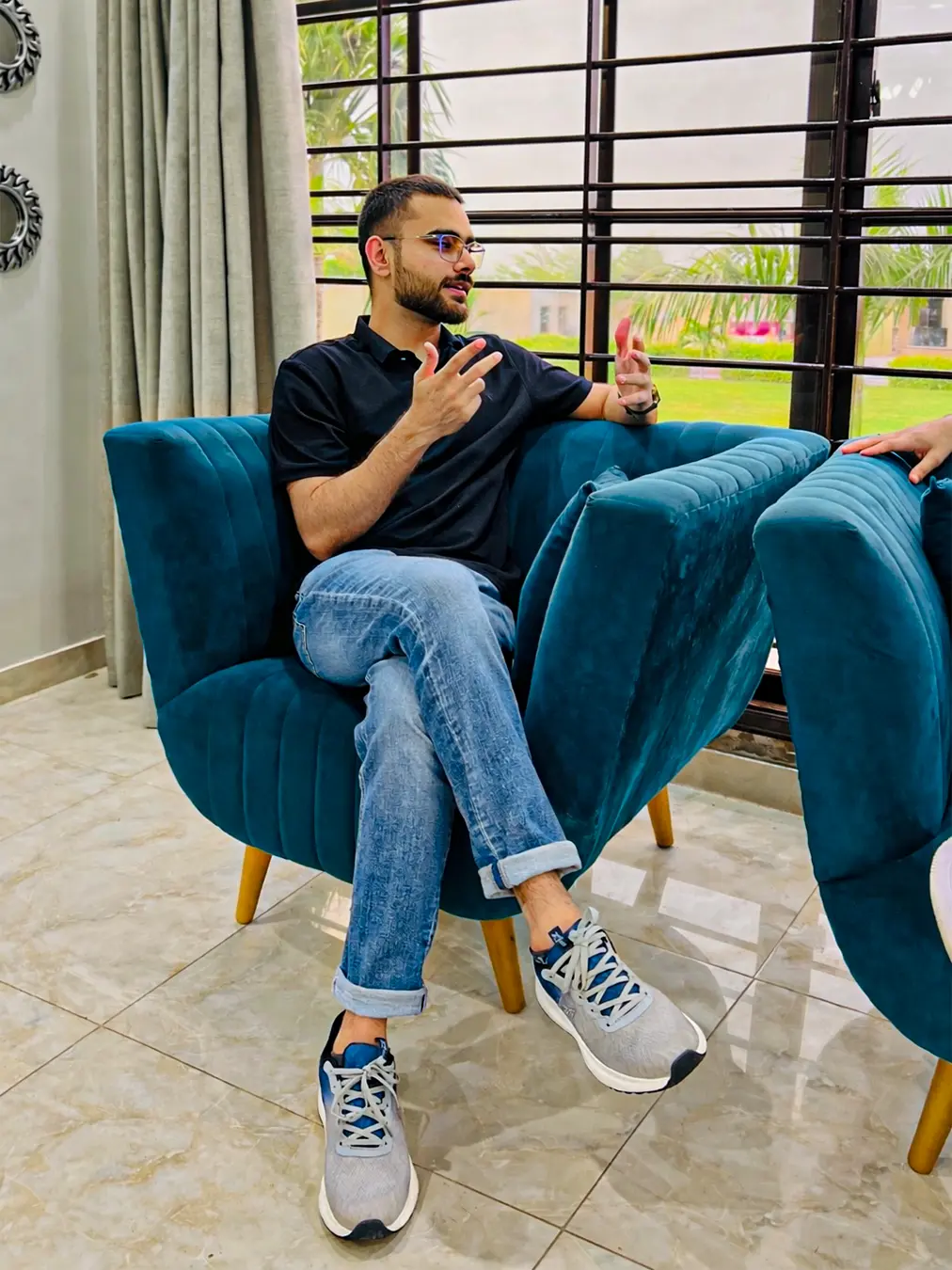 A modern office or living room portrait featuring Abdul Basit RK in a black polo and blue jeans, sitting and gesturing comfortably on a blue velvet armchair, part of a balanced work-life balance triptych with a navy background.